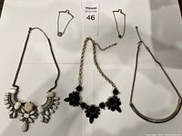 Top view photo showing the five necklaces arranged on a white background, including large geometric pendant necklace, black statement necklace, curved silver necklace, and two delicate pendant necklaces.