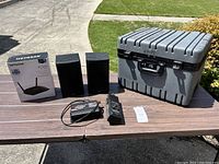 Wide photo showing entire lot including media case, router box, speakers, power converter, and walkie-talkie on outdoor table.