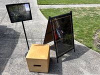 Photo showing all three items together on pavement outside: metal sign stand with black framed sign, metal A-frame sign stand, and wooden storage box.