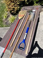 Overview of yard tools and broom laid out on a table outdoors showing broom, shovel, hoe, and rake with various handle colors and metal working ends.