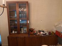 Wide view showing full wood buffet with multiple doors and the glass front cabinet on top containing several glass shelves with décor.