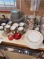 Wide angle showing multiple stacks of white plates with gold patterns, coffee cups, floral coffee pot, floral pitcher, and red glass pieces on kitchen counter