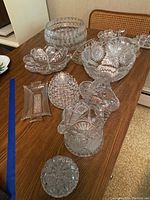 Overview of multiple cut crystal glassware items on wooden table, including bowls, a pitcher, candy dish, trinket box, and other vessels.