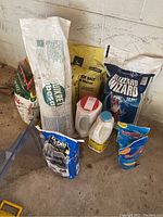 Photo showing multiple household chemical bags and containers including Blizzard Wizard ice melt, rock salt, kitty litter, Damp Rid, and other items stored against a wall on a concrete floor.