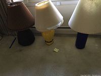 Photo showing all three ceramic lamps with pleated shades aligned on floor near window. Shades show signs of staining.