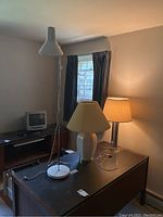 White metal adjustable floor lamp with conical shade, ceramic table lamp with pleated shade, and metal table lamp with beige fabric shade sitting on a dark wooden desk.
