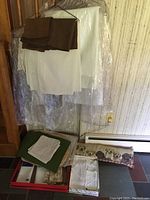 White and brown linens hanging on a stand with a box of folded linens including napkins and placemats on the floor below