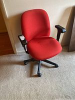 Front and angled views of the red office chair showing cushioned red fabric upholstery, black adjustable armrests, and black caster wheel base.