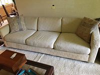 Full view of the beige three-seater upholstered sofa with three removable seat cushions and two decorative throw pillows. The sofa is positioned against a wall and placed on carpeted floor.