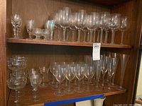 Two wooden shelves displaying various types of clear stemware including wine glasses, champagne flutes, and cordials, some with etched designs.