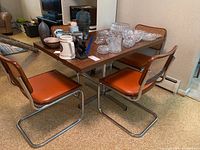 View of the dining table with four chairs surrounding it, table showing faux wood finish and chrome legs, chairs with burnt orange seats and cane backs.