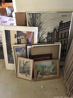 Overview of six framed artworks in various sizes leaning against a wall. Some show urban scenes, others abstract art. Frames are various styles and colors.