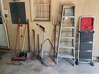 Photo of various yard tools including broom, shovel, pitchfork, weed trimmer, and step ladders against garage wall