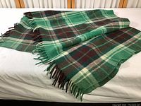 Green brown cream plaid wool blanket with fringed edges laid flat on a bed.