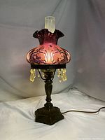 Full view of the lit Fenton lamp showing purple iridescent ruffled glass shade on ornate metal base with hanging crystals.