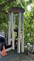 Wind chimes with six silver metal rods hanging from a weathered round wooden disk attached with black cords. A wooden center clapper is hanging inside the rods. The chimes are suspended from a looped black cord and are shown hanging outdoors from a tree branch.