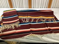 Photo of two folded vintage Mexican serape blankets showing multicolor horizontal stripes with fringed edges and unfinished sides.