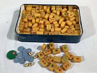 Overall view of miniature ivory-colored Mah Jong tiles with various Chinese characters and symbols in a blue metal tin, along with blue circular tokens, two dice, and one green token outside the tin on white surface.