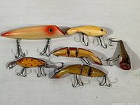 Top-down view of six vintage wood fishing lures arranged on white surface, showing variety in shapes and colors.