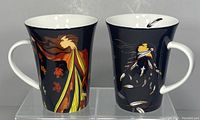 Two ceramic mugs with artistic autumn-themed designs by Maxine Noel on a black background, showing front views.