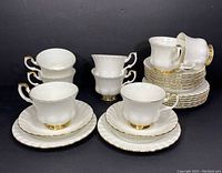 Full set of 8 teacups, 8 saucers and 8 dessert plates arranged grouped and stacked showing gold detailing and scalloped edges.