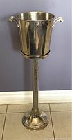 Front view of stainless steel ice bucket mounted on tall pedestal stand, showing overall shape and reflective finish.