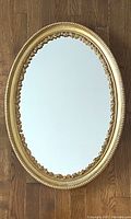 Full view of the vintage oval mirror with gold gilt frame on wood floor showing overall shape and design.