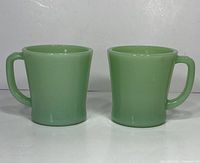 Front view of two jadeite green Fire King mugs side by side on white surface