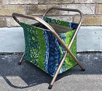 Side view of the folding sewing basket showing tubular metal frame and colorful fabric with green and blue patterns.