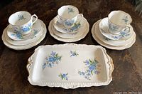 Full set arranged on table showing 3 cups with saucers, piled dessert and salad plates and serving tray with blue Forget Me Not floral pattern and gold accents