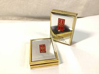 Closed gold-tone compact case with reflective lid and Elizabeth Arden red logo, alongside product box.