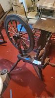 Side view of antique wooden spinning wheel showing large circular wheel with spokes, mounted on turned legs, with attached table surface nearby.