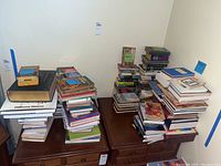 Multiple stacks of various books on wooden dressers against wall, featuring different genres and authors as described by the seller.