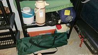 Photo showing multiple camping items: a green camping chair bag, coolers, water dispensers, and sleeping bags on shelving unit and floor.