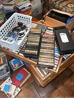 Close-up photo of music cassette tapes arranged on a table with two plastic storage cases and a white basket with cables and headphones.