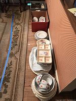 Porcelain plates, serving dishes, cups, trays arranged on floor and inside crate, showing overall quantity and variety.