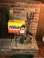 Fireplace tool set with stand, box of Duraflame Firestart, and vintage log roller placed on a brick hearth.