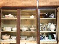 Wide view of wooden cabinet shelves filled with assorted porcelain serveware including covered dishes, teapots, pitchers, mugs, and bowls in various patterns and styles.