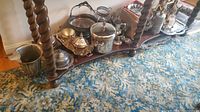 Overview of the silverplate serving pieces arranged on a wooden two-tier stand with twisted supports on a patterned blue carpet.