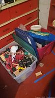 Top angled photo showing two plastic storage bins filled with various toys in a basement room with red cabinetry in background.