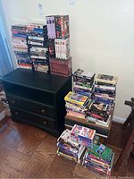 Wide view of the black wooden dresser with drawers holding multiple stacks of various VHS tapes piled high on top and on the floor to the side.