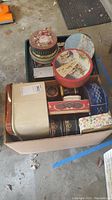 Box containing various vintage metal tins and a wrap dispenser, showing assortment of shapes and colors.