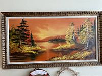 Framed painting showing a wildlife landscape at sunset with orange sky, lush pine trees, and water reflecting the sky, signed by artist Thompson.