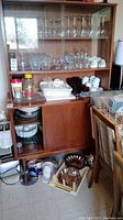 Wooden shelving unit with upper shelves holding various clear glasses including wine glasses, beer glasses, and Luminarc dessert glasses. Middle shelf and counter with coffee cups, jars, and other kitchen items. Lower shelves contain stacked baking pans and bowls. Floor in front shows trays and kitchen containers.