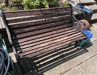 Full side view of metal framed yard bench with wooden slat seat and sculpted metal armrests, placed on concrete patio outdoors