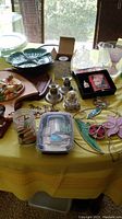 Wide angle photo showing multiple ceramic plates and bowls, pottery dish, glass butter dish, wooden boards, metal candle holders, snow globe, and boxed collector spoons arranged on a yellow tablecloth.