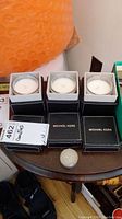 Three Michael Kors candles in small clear glass holders on a brown table. Each candle is paired with a black Michael Kors branded box with white lettering.
