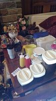 Full lot view showing travel mugs, kitchen canisters, condiment bottles, faux flowers, vases, and other kitchen containers arranged on a wooden table.