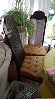 Two wood chairs with cane backs and tufted mustard yellow seats seen from side, showing carved wood tops and seats next to window with plants.