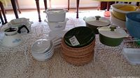 Overview of vintage Corning Ware tea pot, casserole dishes, skillets, and additional kitchenware on white display rack on carpeted floor under table.